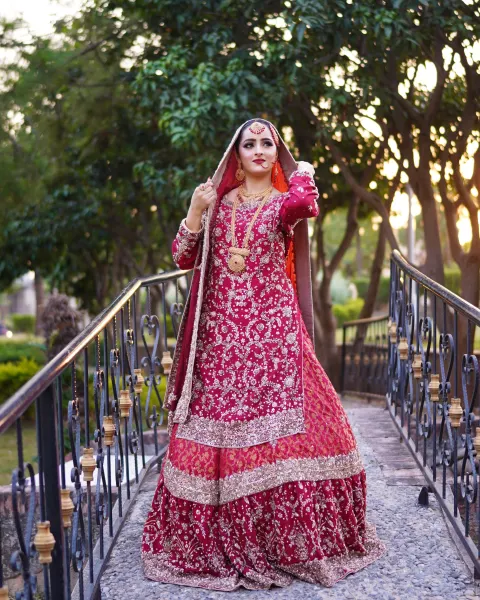 A Pakistani bride in a heavily embroidered crimson red and maroon lehenga kurti set posing on a stone bridge with iron railings, captured by AM Productions.