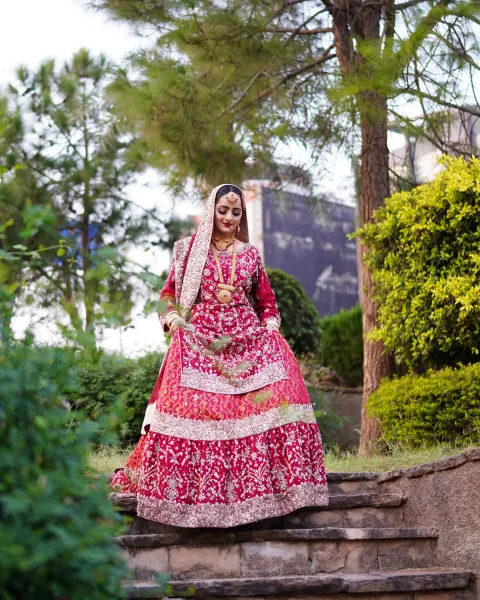 A Pakistani bride looking down gracefully while walking down rustic stone stairs in a heavily embellished crimson red and maroon lehenga, captured by AM Productions.