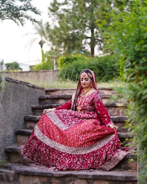 A Pakistani bride sitting gracefully on wide rustic stone stairs, wearing a heavily embroidered crimson red and maroon lehenga while looking down thoughtfully, captured by AM Produ