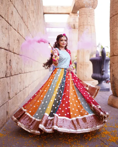 A Pakistani Mehndi bride twirling in a colorful multi-paneled lehenga and light blue choli while holding a pink smoke bomb near an ancient Egyptian-themed stone wall, shot by AM Pr