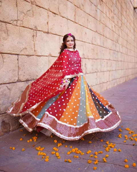 A beautiful Pakistani Mehndi bride looking over her shoulder while twirling in a colorful paneled lehenga and a red chunri-style dupatta against a large sand-colored stone block wa