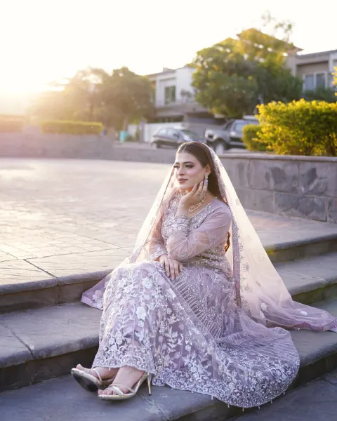 A Pakistani bride sitting on outdoor stone steps in a pastel lilac and silver embroidered pishwas dress during golden hour, captured by AM Productions.