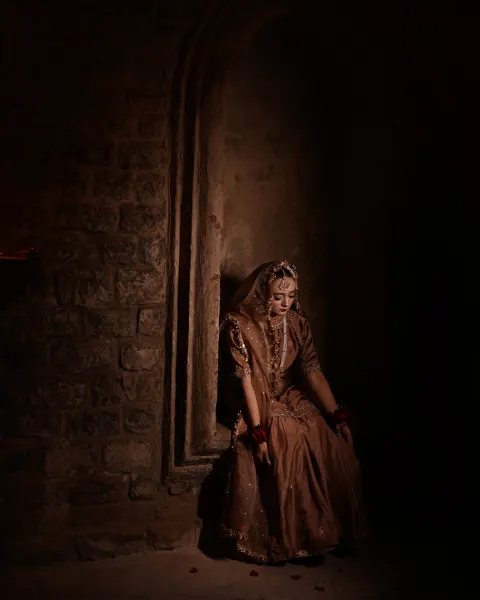A Pakistani bride sitting in a dimly lit rustic stone archway alcove looking down, wearing a metallic mocha-brown gharara with deep red roses on her wrists, captured by AM Producti