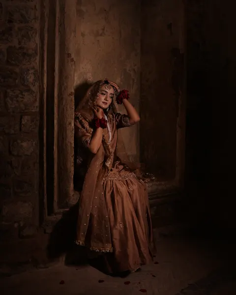 A Pakistani bride posing elegantly with her hands near her head while sitting in a moody, dimly lit rustic stone archway alcove, captured by AM Productions.