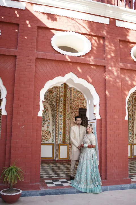A couple in traditional Pakistani wedding attire posing inside a historic red brick archway, captured by AM Productions.