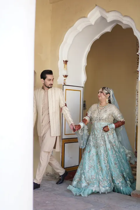 A newlywed couple in traditional Pakistani wedding attire holding hands while walking through a classic archway, captured by AM Productions.