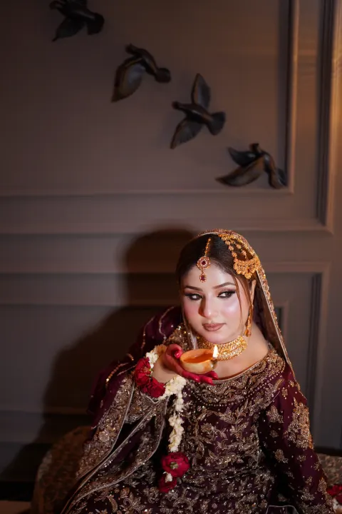 A gorgeous South Asian bride in a deeply embroidered maroon and gold bridal outfit, holding a lit diya while adorned with traditional gold and floral jewelry — Islamabad & Rawalpindi photography by AM Productions.