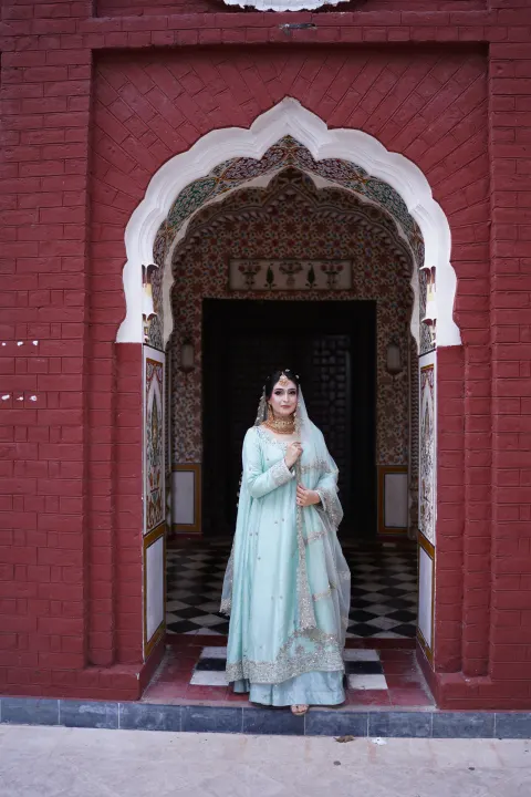 Traditional Pakistani bridal portrait featuring a bride in a mint green pishwas standing under a historic red brick archway, captured by AM Productions in Islamabad and Rawalpindi.