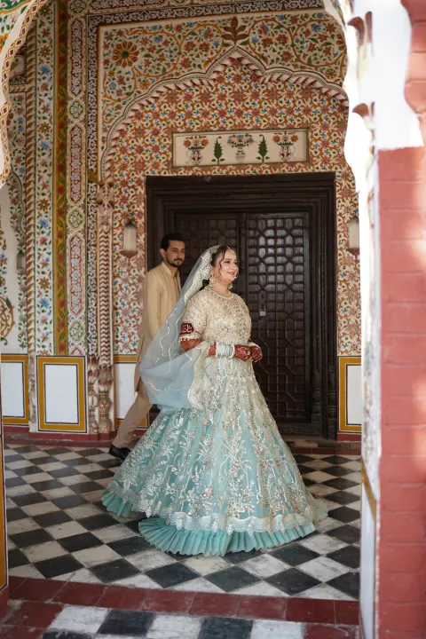 A Pakistani bride in a heavily embellished pastel mint green and gold lehenga smiling during a wedding shoot by AM Productions at a historic haveli with intricate fresco walls.