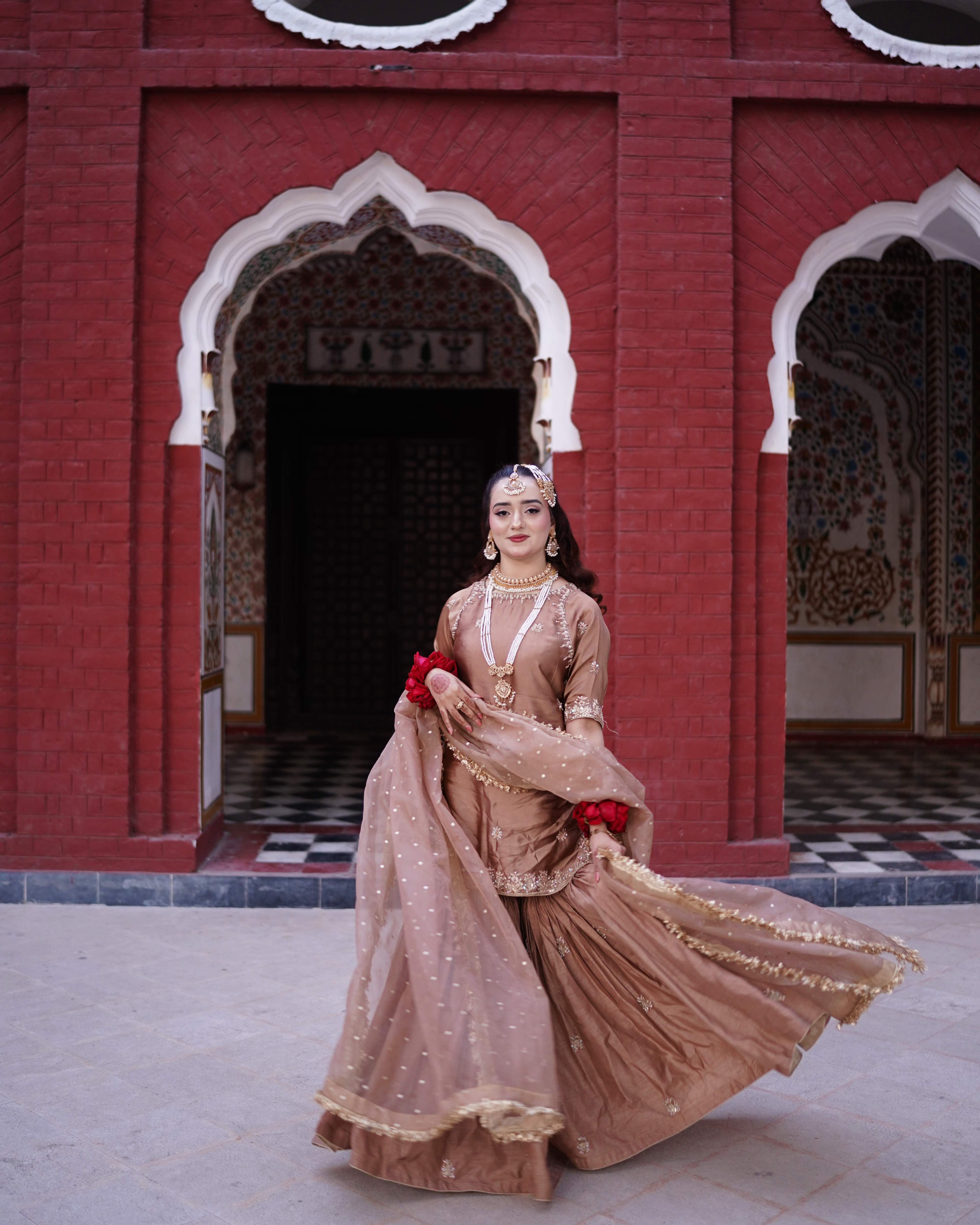 A Pakistani bride gracefully twirling her metallic mocha-brown gharara in front of a grand red brick heritage building with traditional Mughal arches, captured by AM Productions.