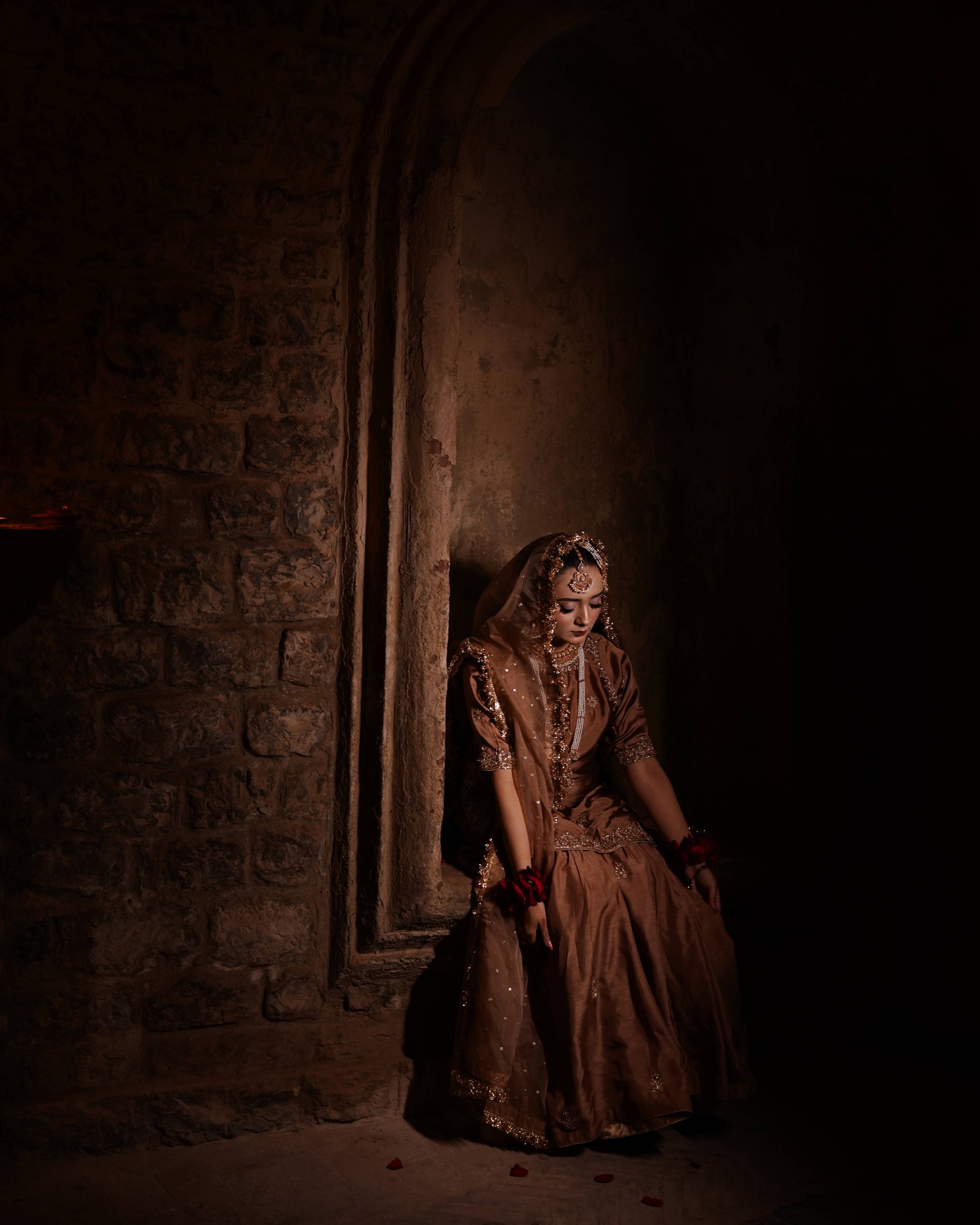 A Pakistani bride sitting in a dimly lit rustic stone archway alcove looking down, wearing a metallic mocha-brown gharara with deep red roses on her wrists, captured by AM Producti