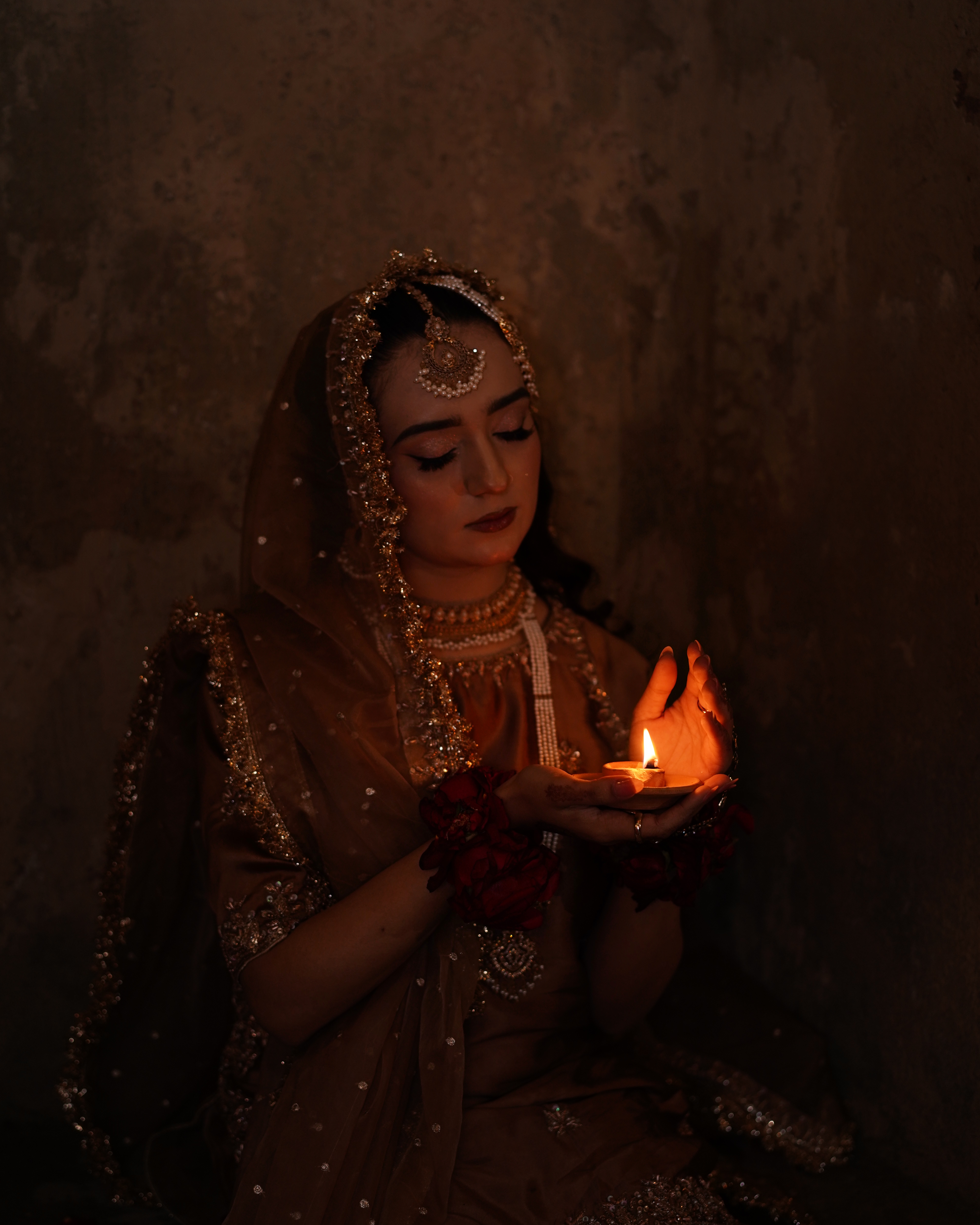 A close-up portrait of a Pakistani bride with closed eyes, gently holding a lit clay lamp (diya) that illuminates her face in a dark, moody setting, captured by AM Productions.