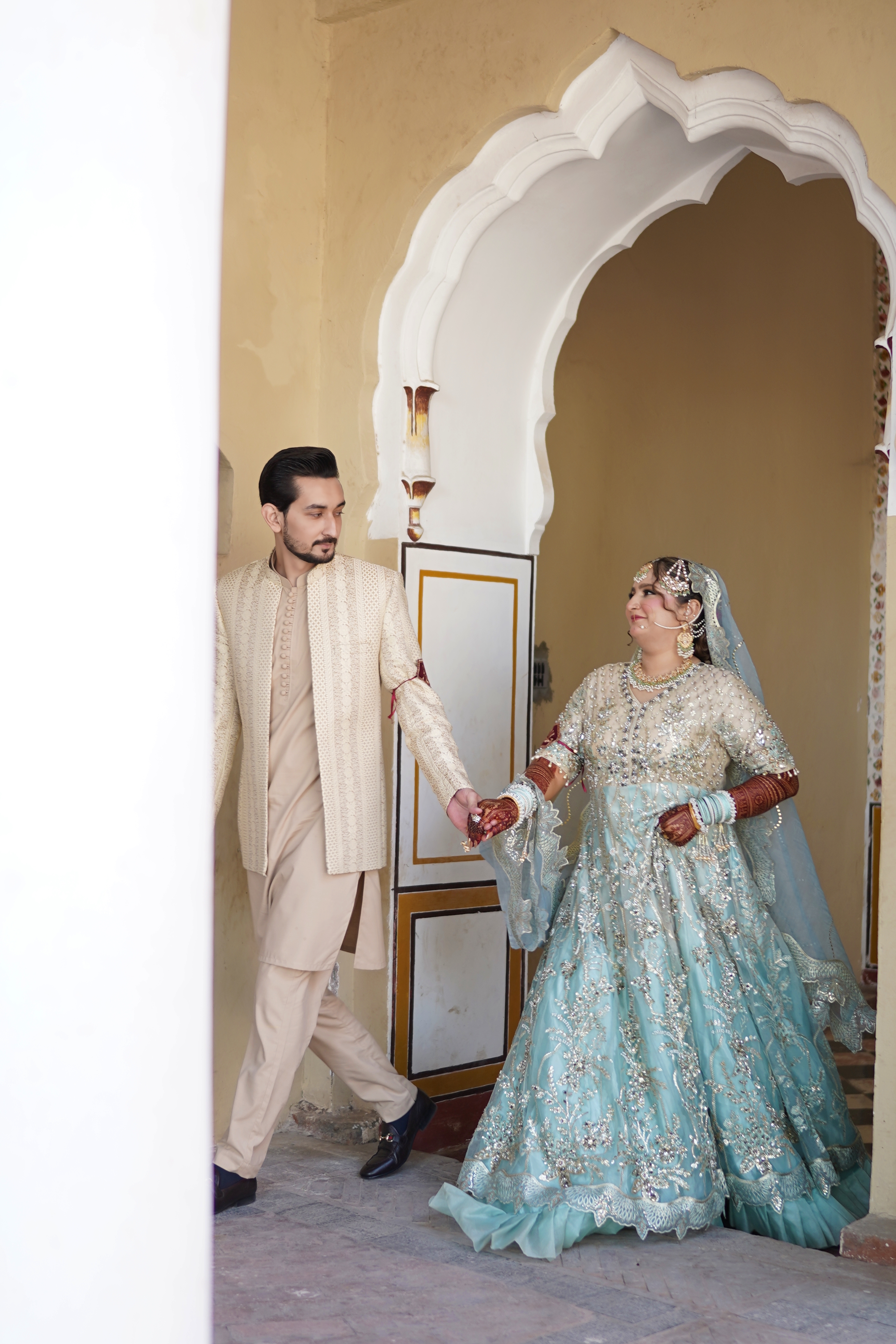 A newlywed couple in traditional Pakistani wedding attire holding hands while walking through a classic archway, captured by AM Productions.