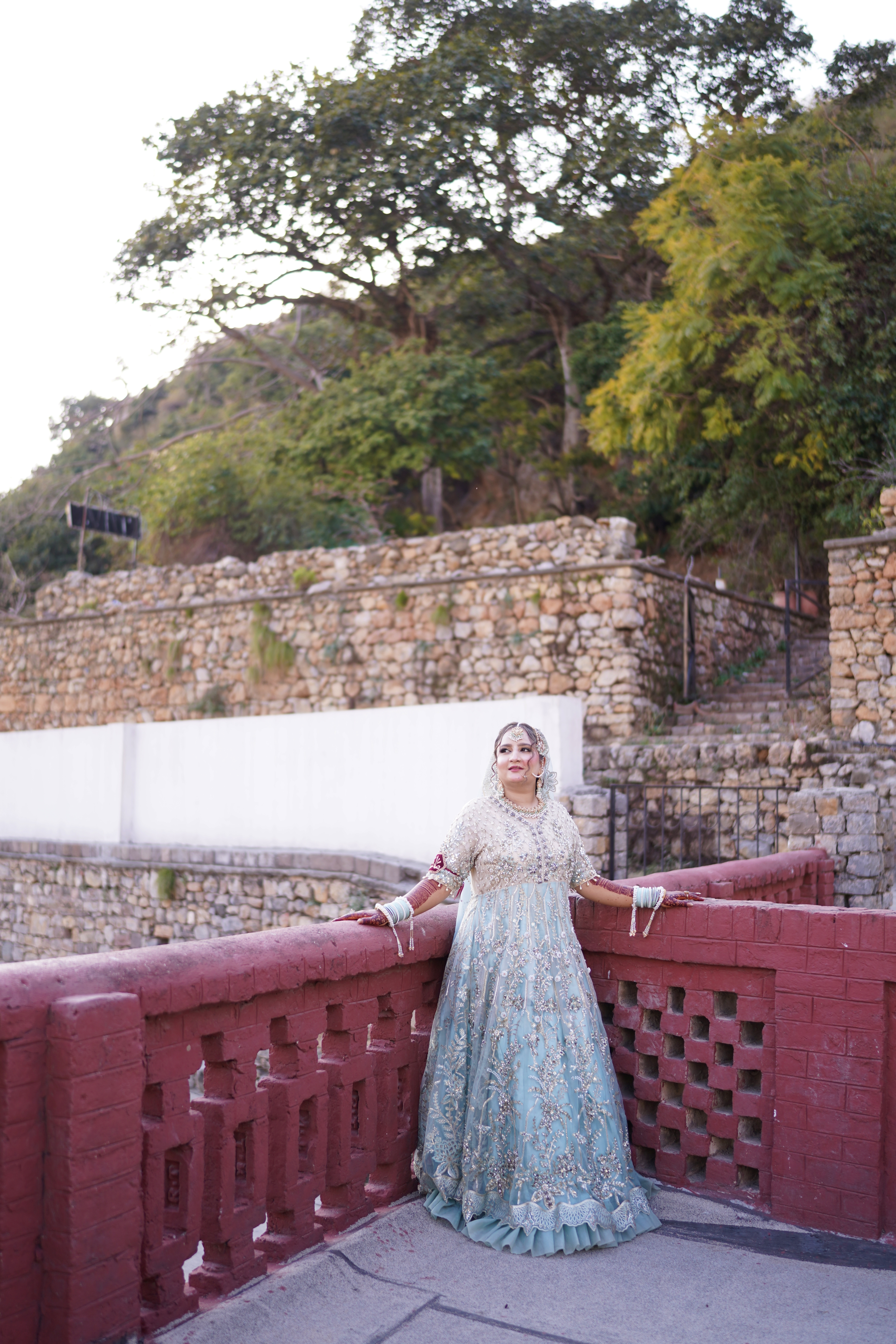 A solo Pakistani bride in a premium pastel mint green and gold embellished lehenga posing gracefully against a terraced stone wall and lush green hillside, featured in a wedding re